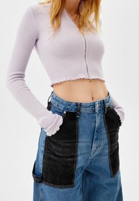 Mujer vestida con un top corto de manga larga acanalado en lila y cierre, y unos jeans de mezclilla de dos tonos, azul y negro, de cintura alta, con las manos en los bolsillos.