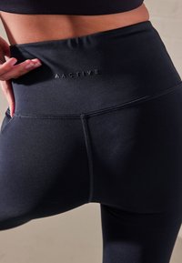 Schwarze, hoch taillierte Sportleggings mit glatter Textur und Nahtdetails, versehen mit einem dezenten Logo am Bund.