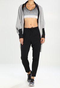 Žena v šedém sportovním podprsence, volné šedé bundě, černých teplákových kalhotách a šedo-černých sportovních botách, stojící na bílém pozadí.