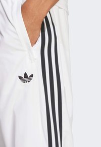 Vitaidrottsbyxor i vitt med svart tre-stripes design och logotyp. Tillverkade av ett mjukt material, med sidofickor och dragkedja.