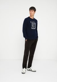 Pull en maille bleu marine avec un design de lettre "D" gris clair, associé à un pantalon marron foncé et des chaussures de sport noir et blanc.