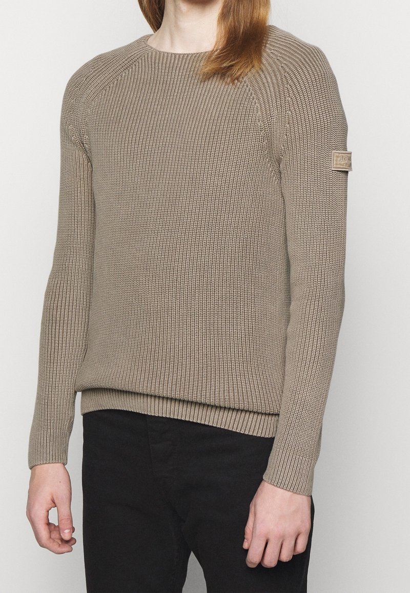 Pull en tricot beige avec texture côtelée, col rond et manches longues. Présente un patch logo sur la manches gauche. Porté avec un pantalon noir.