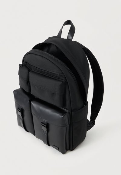 Sac à dos noir avec deux poches avant à rabat équipées de fermetures à boucle et un compartiment supérieur zippé, présenté sur un fond clair uni.