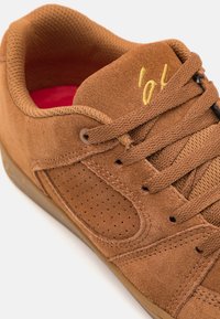 Baskets en daim marron avec panneau latéral perforé, lacets assortis, logo jaune brodé sur la languette et semelle intérieure rouge.