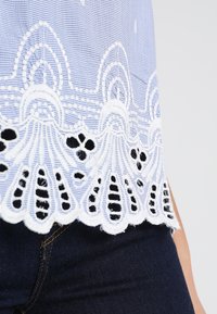Tela a cuadros azul y blanco con intrincado bordado de encaje blanco que presenta patrones recortados a lo largo del dobladillo. Jeans oscuros abajo.