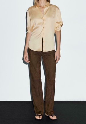 Cream satin button-up shirt with short sleeves, paired with brown suede straight-leg pants. Light necklace completes the look.