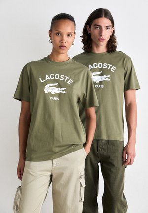 Olive spalvos medvilniniai marškinėliai su balta Lacoste logo ir krokodilo grafika; modelis vilki béžinius krovinių kelnes; rodomi ant dviejų figūrų.