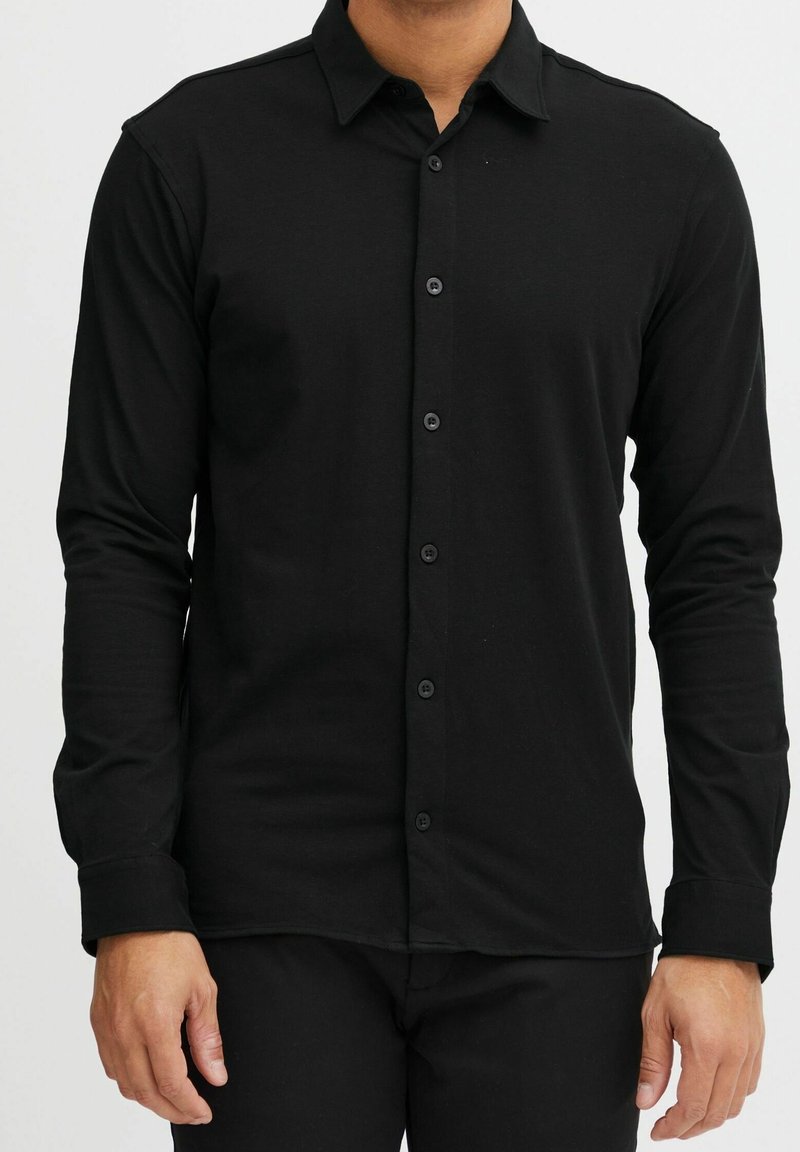 Homme portant une chemise noire unie à manches longues avec col et un pantalon noir, debout devant un fond blanc.
