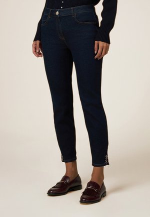 Jeans de mezclilla azul marino con un corte slim, que presentan bolsillos frontales, costuras a tono y puños adornados. Combinados con mocasines de color burdeos oscuro.