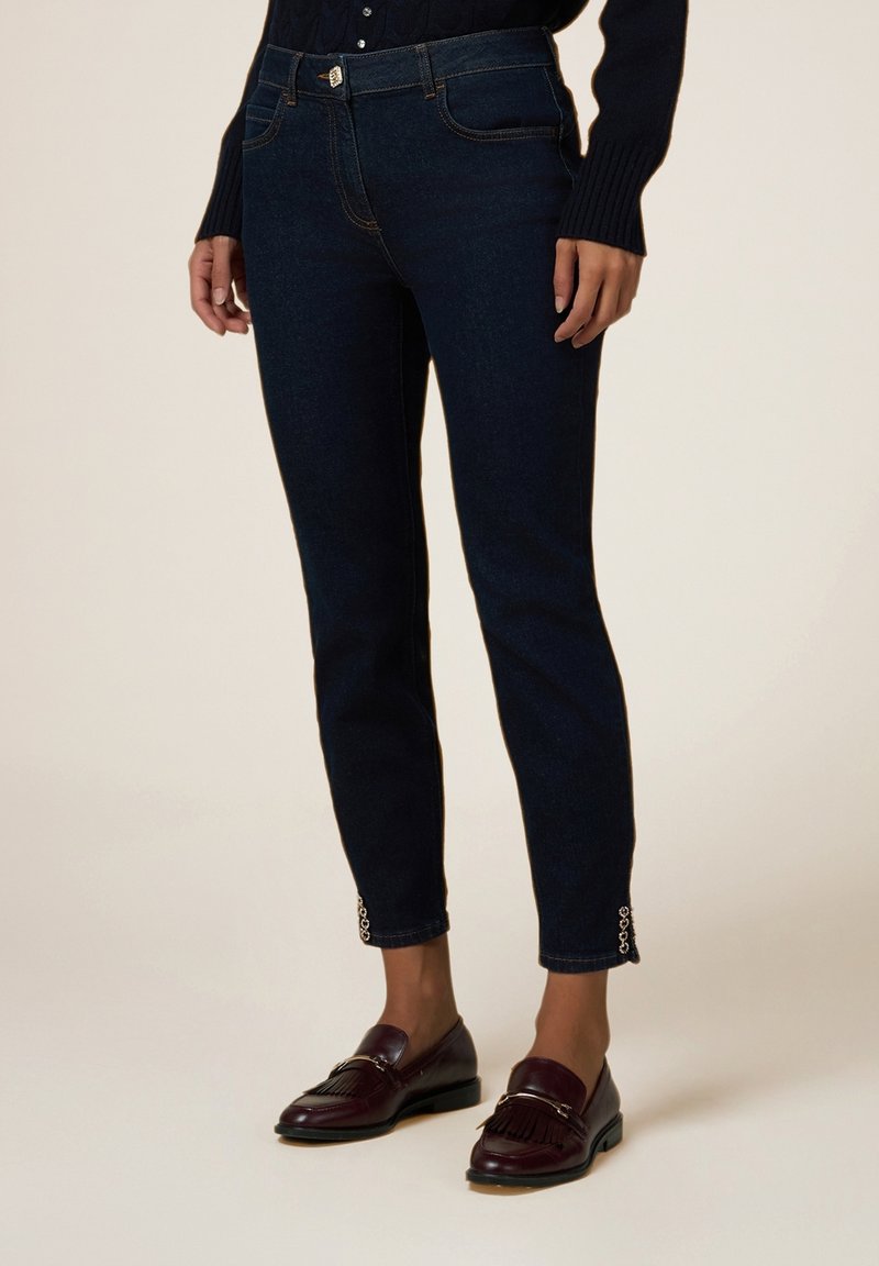 Jeans de mezclilla azul marino con un corte slim, que presentan bolsillos frontales, costuras a tono y puños adornados. Combinados con mocasines de color burdeos oscuro.