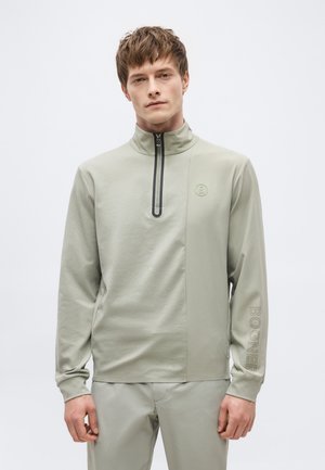 Giovane uomo che indossa una felpa con zip grigio chiaro con collo alto e pantaloni abbinati, in piedi davanti a uno sfondo semplice.