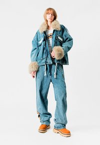 Chaqueta de mezclilla azul de gran tamaño con forro de piel, combinada con pantalones de mezclilla azul holgados y calzado naranja. Presenta texturas distintas y un contraste de colores.