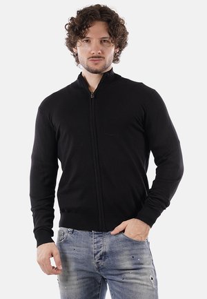 Uomo con capelli ricci che indossa un maglione zip-up nero e jeans blu consumati, in piedi con una mano in tasca su uno sfondo semplice.