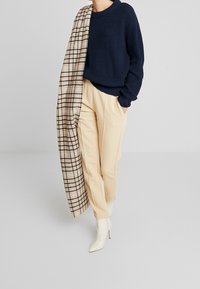Maglione lavorato a maglia blu navy, pantaloni beige plissettati e stivaletti bianchi a punta alla caviglia, accentuati da una sciarpa beige a scacchi adagiata sulla spalla.