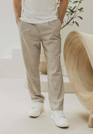 Homme portant un pantalon beige et des baskets blanches, debout à l'intérieur à côté d'une chaise en rotin courbée et d'une plante en pot.