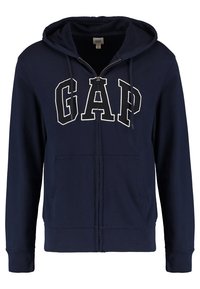Mörkblå zip-up huvtröja med stora vita "GAP" bokstäver, framfickor och snörning i huvan. Tillverkad av mjukt bomullstyg.