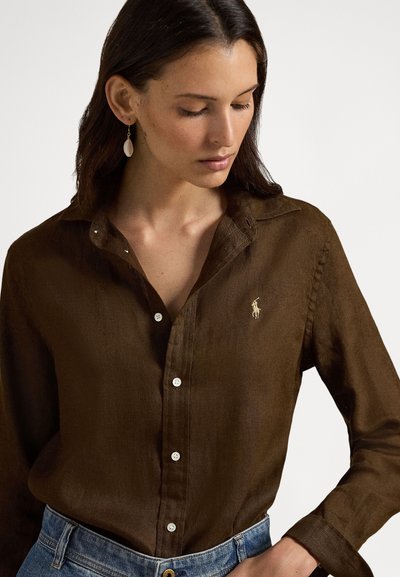 Brūna linu pogājama krekls ar klasisku apkakli, kas ir ar nelielu izšūtu logotipu un baltām pogām. Valkāts kopā ar zilām džinsa biksēm.