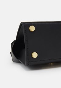 Fondo di una borsa in pelle nera con due piedini rotondi in metallo dorato visibili su uno sfondo bianco.