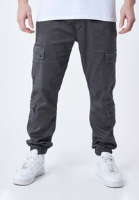 Pantalones cargo grises con múltiples bolsillos laterales, puños elásticos y un acabado de tela suave, combinados con zapatillas blancas.