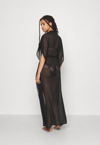 Svart gjennomsiktig kaftan med korte ermer, som har en bred, åpen design. Stoffet er lett og litt teksturert, og viser en blomstermotiv under.