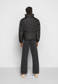Giacca a vento nera con colletto alto e sezioni trapuntate, indossata sopra jeans in denim scuri a gamba larga, con capelli intrecciati visibili.