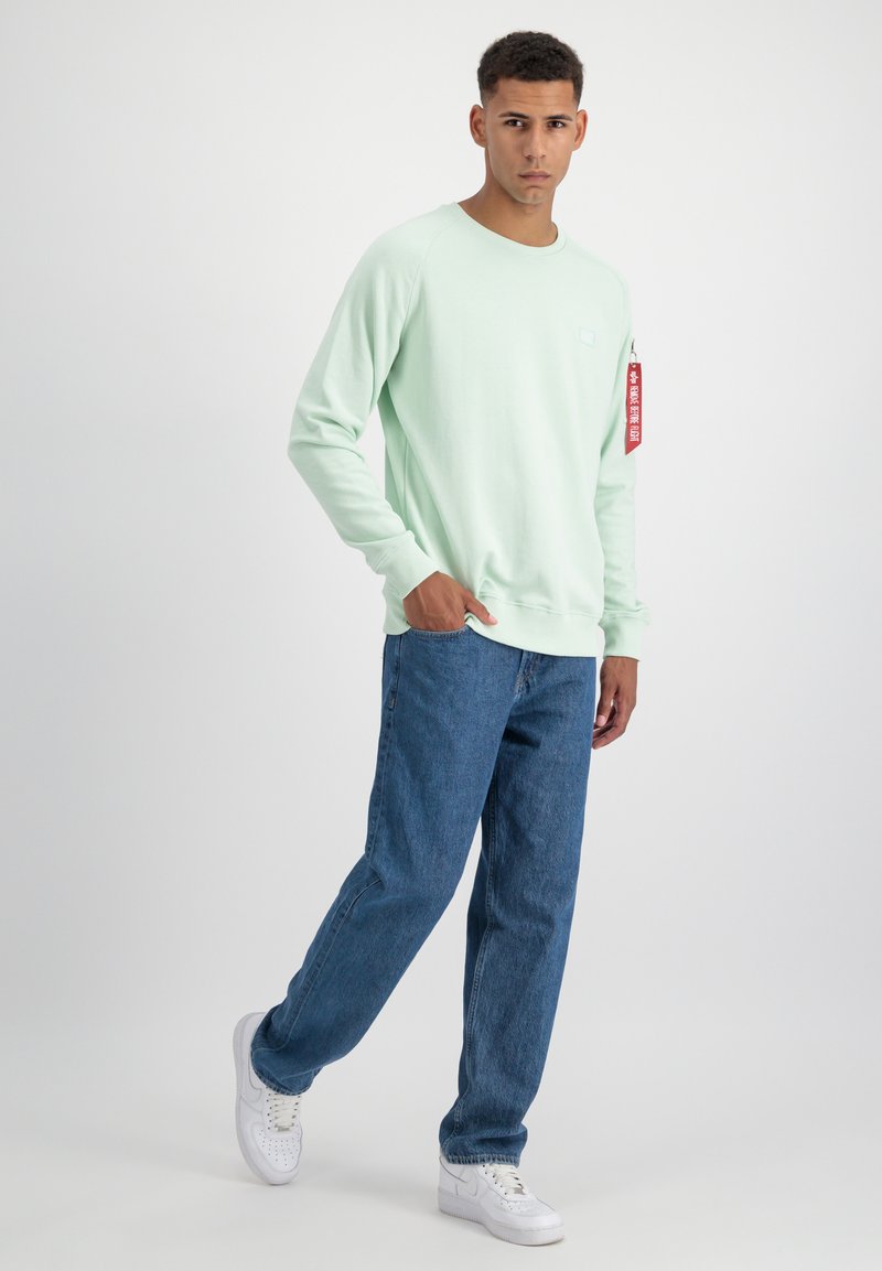 Jeune homme portant un sweat-shirt vert clair avec une étiquette rouge sur la manche, un jean bleu et des baskets blanches, debout sur un fond blanc uni.