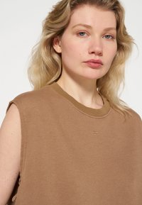 Bruin mouwloos topje van zacht materiaal, met een ronde halslijn en rafelige randen met subtiele stikdetails.