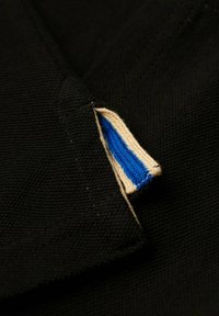 Tessuto nero con un piccolo etichetta caratterizzata da una striscia blu e un bordo beige, cucita nella cucitura, che mostra dettagli sulla texture e sul materiale.