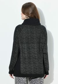 Mujer con cabello castaño que lleva un suéter moteado en negro y gris y pantalones gris con estampado de leopardo, de espaldas frente a una pared blanca.