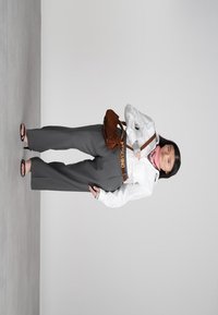 Mlada ženska v beli bluzi in sivih širokih hlačah stoji, drži rjav nahrbtnik, nosi rjav pas in črne sandale z odprtim prstom pred sivo ozadje.