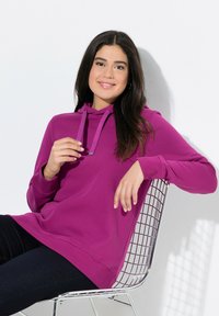 Felpa viola brillante con tasca a marsupio, cappuccio con cordino e polsini a costina, abbinata a jeans scuri, indossata mentre si è seduti su una sedia in rete.