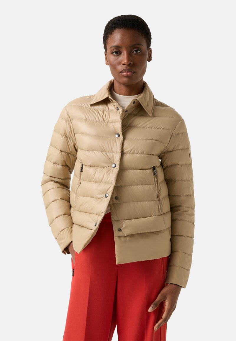 Beżowa kurtka puchowa z poziomym pikowaniem, z zapięciem na zatrzaski, dwoma bocznymi kieszeniami i kołnierzem koszulowym. Noszona na czerwonych spodniach, podkreślając kontrast.