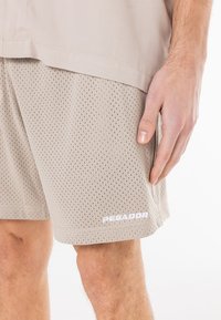 Shorts de sport beiges en tissu en maille, avec une coupe ample, de petites poches latérales et un logo "PEGADOR" blanc sur le bas à gauche.