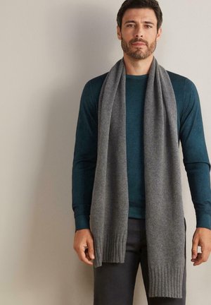 Scaldino grigio a maglia con estremità a coste, appoggiato attorno al collo di una persona che indossa una maglietta a maniche lunghe verde acqua e pantaloni scuri. Texture liscia.