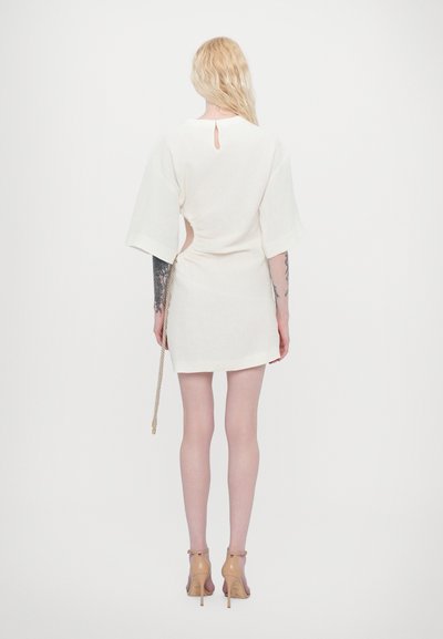White short dress with a back keyhole and side gather detail, featuring wide sleeves and an attached rope belt, paired with clear heels.