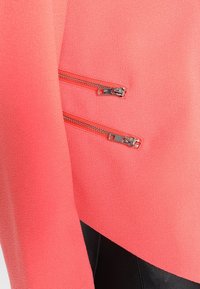 Veste corail en tissu texturé, avec deux poches zippées argentées à l'avant. Le design comprend une section contrastante noire à la base.