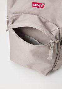 Šedý látkový batoh s predným vreckom, ktorého zips je zakrivený a nad ním sa nachádza červené logo Levi's. Materiál vyzerá textúrovaný a odolný.