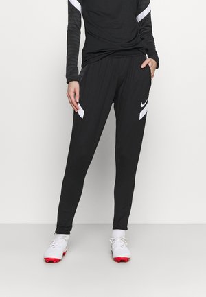 Personne portant un pantalon de sport noir avec des logos Nike blancs et des chaussures de football blanches avec des crampons rouges, debout devant un fond uni.