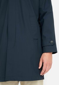 Giacca impermeabile blu navy dal taglio dritto, con tasca laterale, polsini con bottoni e texture liscia. Design minimalista con linee pulite.