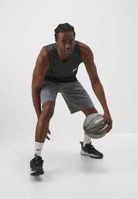 Jugador de baloncesto con una camiseta sin mangas gris y pantalones cortos grises, botando un balón de baloncesto texturizado, llevando zapatillas negras y calcetines blancos con un logo.