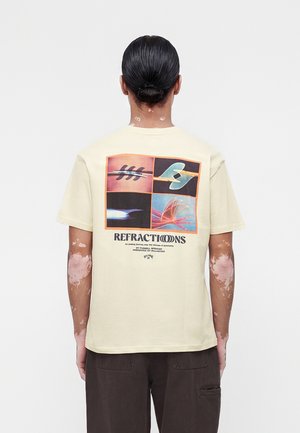 Άτομο με μαλλιά δεμένα πίσω φοράει κρεμ μπλουζάκι με πολύχρωμα αφηρημένα τετράγωνα σχέδια και το κείμενο "REFRACTIONS" στην πλάτη.
