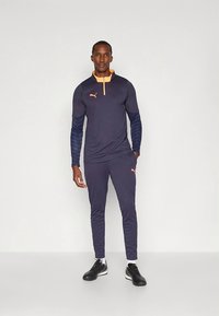 Sötétkék sport melegítő hosszú ujjakkal, narancssárga gallér részlettel és texturált kék ujjakkal. Karcsúsított szabással és fekete sportcipővel.