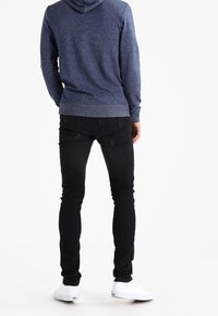 Felpa con cappuccio blu e texture, con tasca a marsupio; abbinata a jeans neri aderenti e sneakers bianche, vista da dietro.