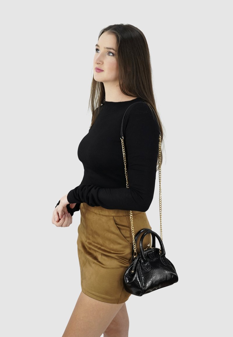 Zwarte top met lange mouwen, tan suède rok en een kleine zwarte handtas met textuur en gouden schouderband. Model staat tegen een grijze achtergrond.