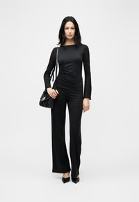 Γυναίκα όρθια, φοράει εφαρμοστή μαύρη μπλούζα με μακριά μανίκια, φαρδιά μαύρη παντελόνια, μαύρες μυτερές γόβες, κρατώντας μαύρη τσάντα ώμου.