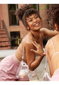 Hellrosa ärmelloses Oberteil und passende hoch taillierte Shorts. Transparente Tasche hält cremefarbene Behälter. Im Hintergrund ist ein Backsteingebäude und eine Treppe zu sehen.