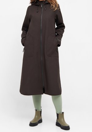 Femme portant un long manteau noir avec capuche, mains dans les poches, leggings verts côtelés et bottines vert olive avec semelles noires.