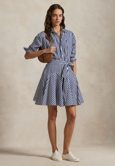 Sieviete stāv, valkājot zilas un baltas vertikālas svītras krekli kleitu ar sasietu jostu, baltas sporta kurpes un tur rokā nelielu brūnu plecu somu.