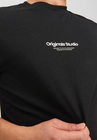 Black cotton t-shirt featuring a round neck and short sleeves. Front print includes "Originals Studio" and additional text in white.