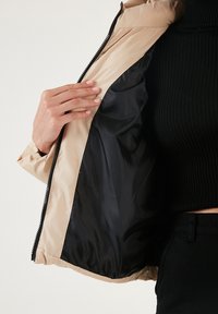 Beige puffer jacket with a black interior, featuring a zip closure and a smooth satin-like texture. The sleeve has a cuff and open hem.